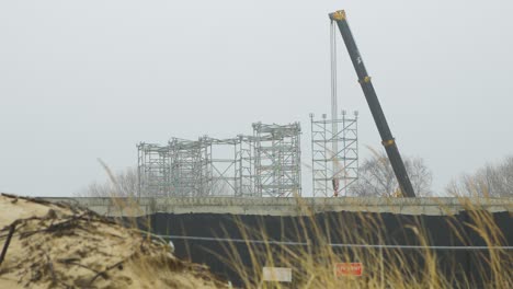 Intensive-work-on-the-construction-site-located-near-the-coast-of-the-sea,-mobile-crane-moving,-white-sand-piles,-overcast-day-with-fog,-distant-medium-shot