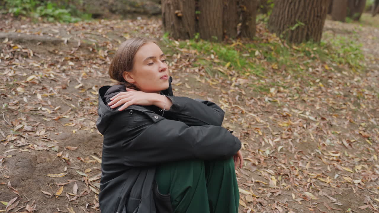 mulher jovem sentada perto de um riacho na floresta de outono, usando jaqueta puffer preta e calças verdes, descansando no chão coberto de folhas, com as mãos cruzadas sob o queixo, olhar reflexivo, luz natural suave, clima tranquilo, close-up