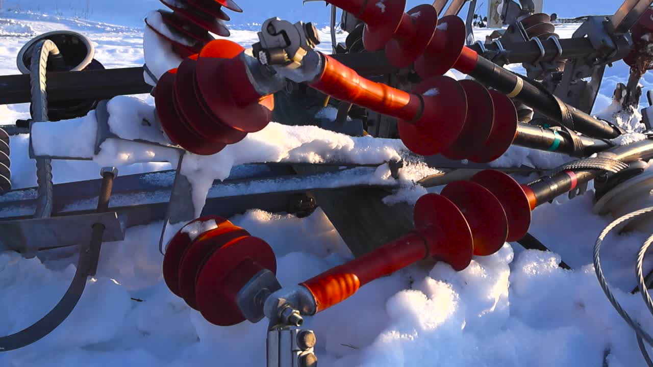 Vista angolata dall'alto di isolatori ferroviari ad alta tensione di colore rosso vivido adagiati nella neve profonda, con ghiaccio e brina illuminati dalla calda luce solare invernale e un lento movimento cinematografico della telecamera.