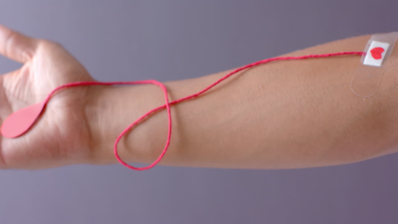 Mano di uomo birazziale con goccia di sangue, filo rosso e cerotto a forma di cuore su sfondo grigio, al rallentatore.