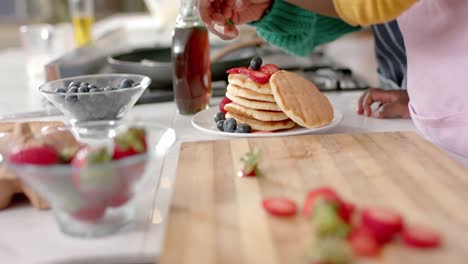 Hands-of-african-american-mother-and-daughter-decorating-pancakes-with-fruits,-slow-motion