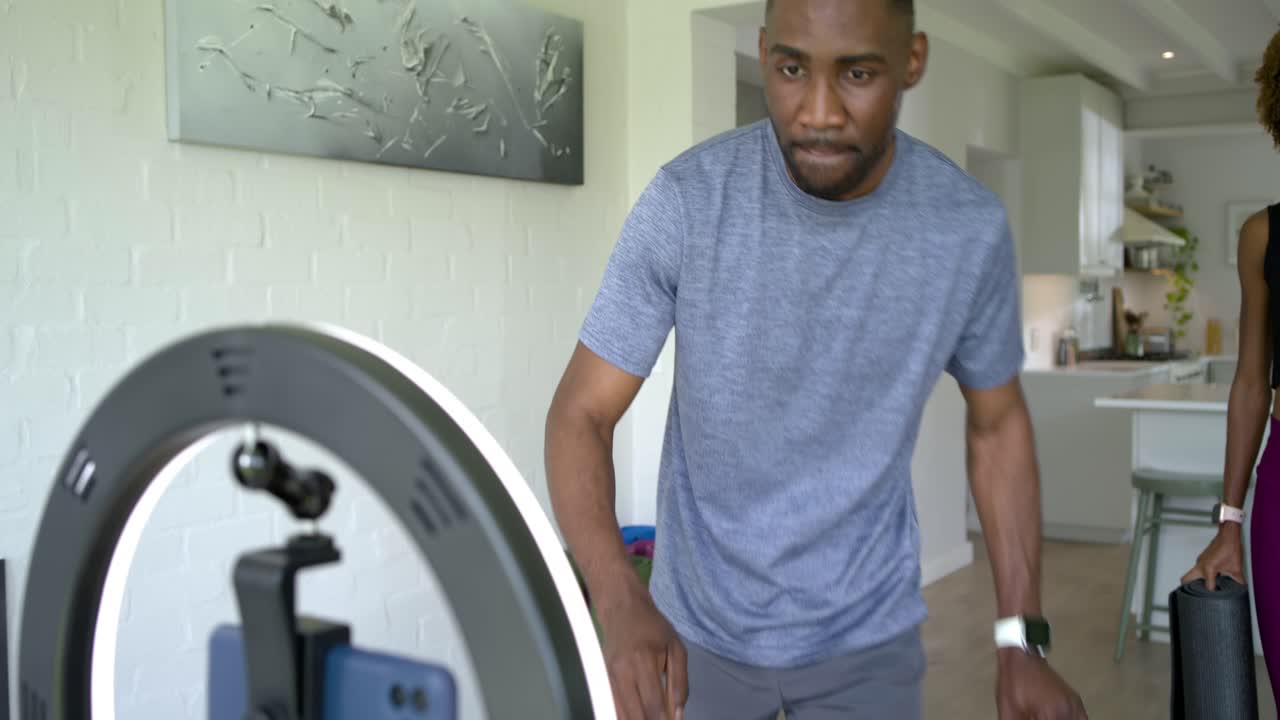 African American couple entering with mats in gymwear, adjusting phone rig, starting workout stream