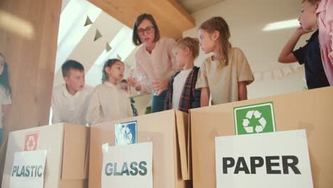 una chica rubia con un peinado bob enseña a los niños en edad preescolar cómo doblar correctamente botellas de plástico y clasificar la basura por tipo de material en un club para preparar a los niños para la escuela