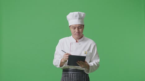 Asian Man Chef Taking Note On A Tablet While Standing In The Green Screen Background Studio