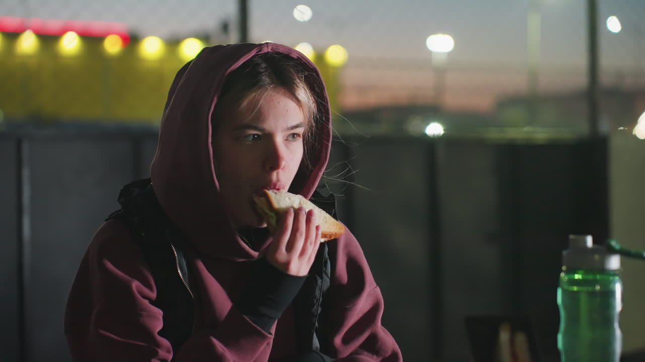 Female athlete sitting outdoors at dusk on bench under bright city lights eating bread while holding water bottle beside sports bag and phone on table after workout enjoying snack rest in urban park