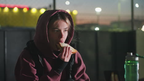 female athlete sitting outdoors at dusk on bench under bright city lights eating bread while holding water bottle beside sports bag and phone on table after workout enjoying snack rest in urban park