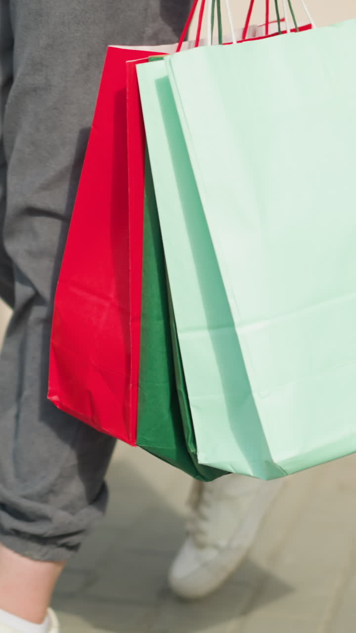 vista de la pierna de alguien sosteniendo bolsas de compras verdes, menta y rojas mientras camina casualmente por un camino entrelazado, las bolsas se balancean con cada paso, y la sombra del caminante es visible en el suelo
