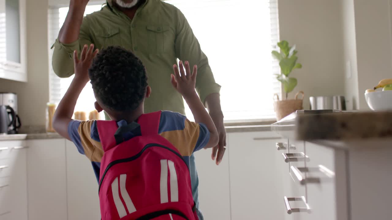 Happy african american grandfather high fiving with grandson in kitchen as he leaves for school