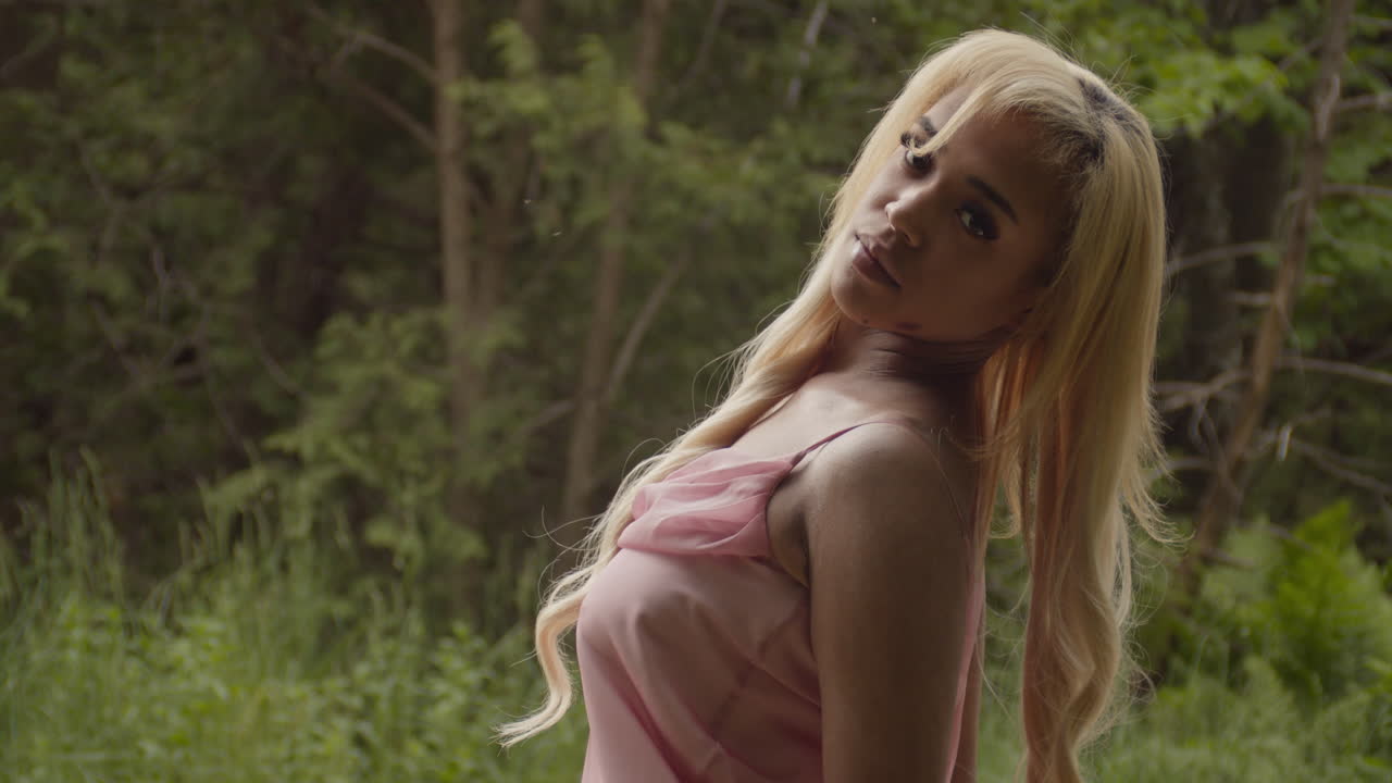 una mujer de color ébano con el cabello largo y rubio, vestida con un vestido rosa, se encuentra a lo largo de un camino de grava en el bosque. de cara lejos de la cámara, gira su torso hacia la cámara, ya que se inclina desde su cintura hasta su cara.