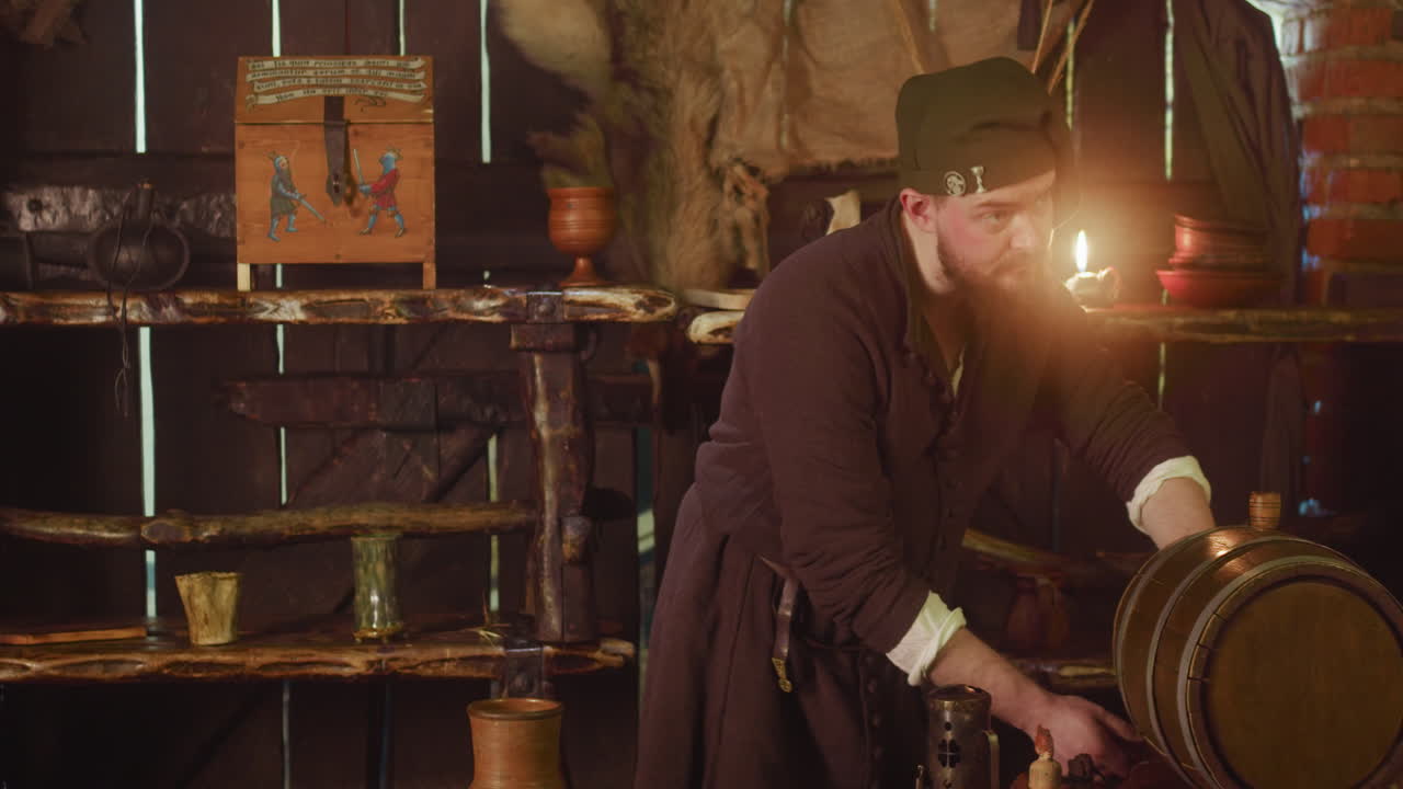 Barkeeper pours ale from barrel and turns to counter at inn. Plump bartender in medieval suit looks with servile smile at guest in tavern. Fantasy world