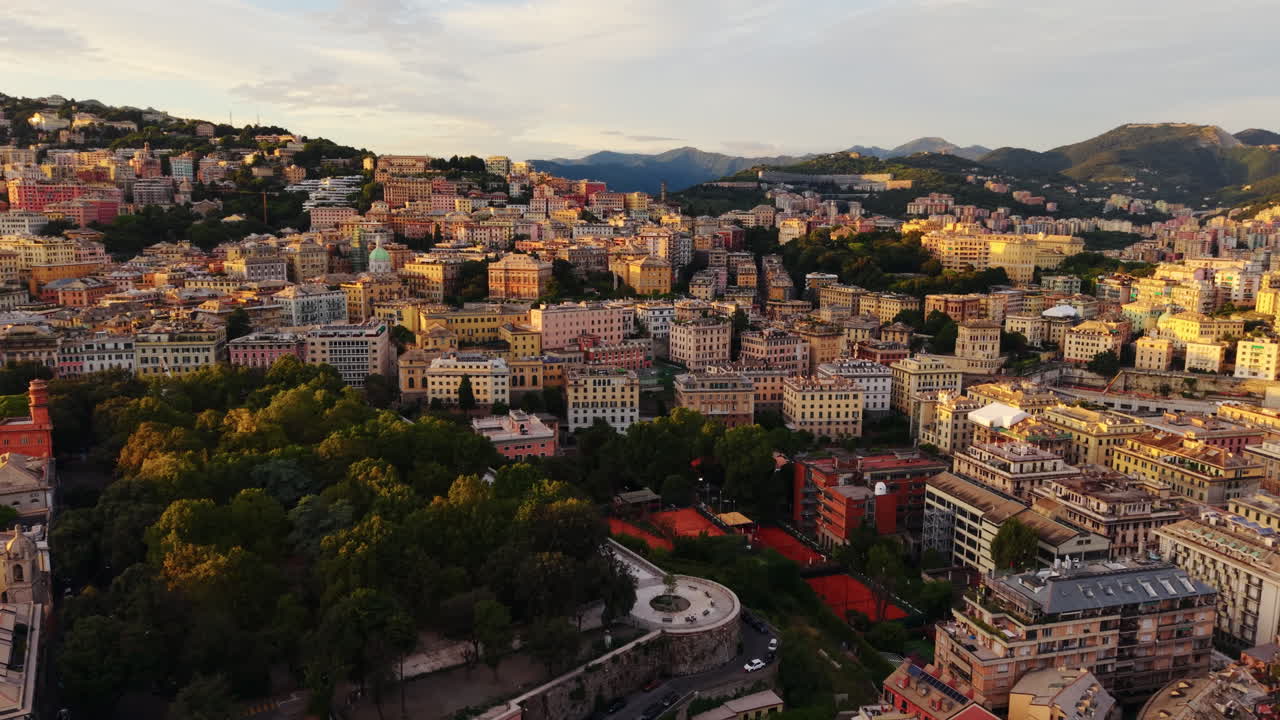Drone panning shot over Genoa old town at sunset, showing dense buildings, a park with trees, tennis courts, and mountains in the background