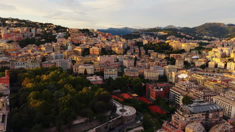 drone panning shot over genoa old town at sunset, showing dense buildings, a park with trees, tennis courts, and mountains in the background