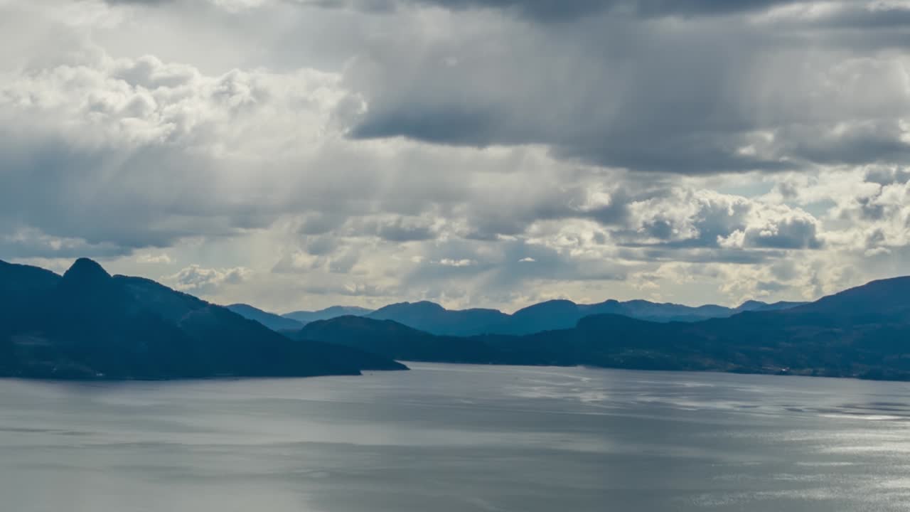 Thick white-gray clouds whirling and moving above the dark blue waters of the Hardanger fjord, Norway. Rays of sun are piercing through the clouds, lighting dark waters. Mountains tower on the horizon
