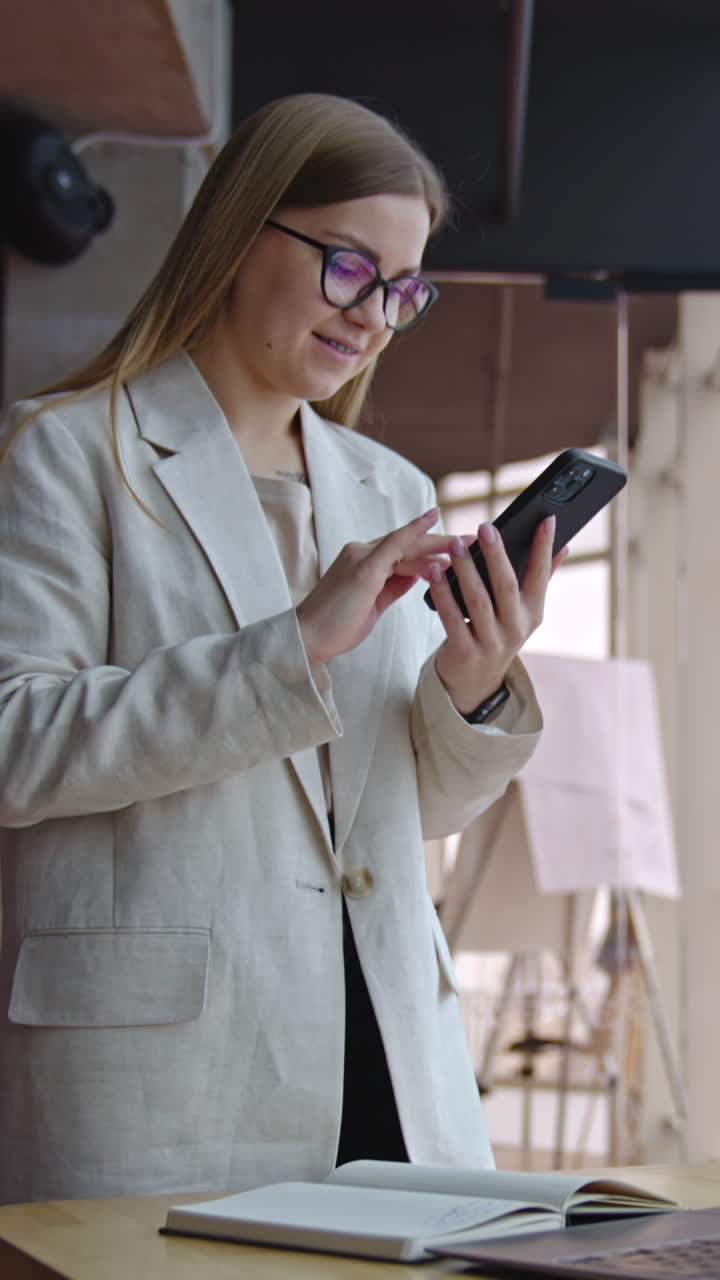 Good-looking lady standing in front of desk and taking notes on paper. Smiling woman makes a phonecall and looks at the screen. Vertical video