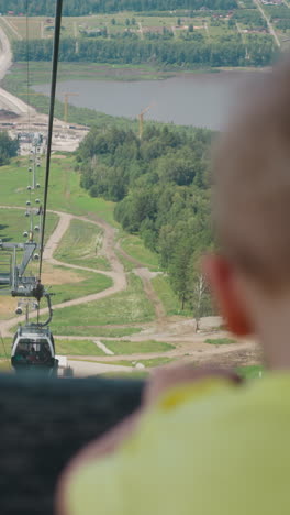 little boy looks at wide green valley with road and lake leaning on rear chair back in gondola of ropeway at resort close backside view slow motion