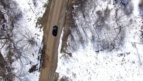 top down aerial drone footage of a black car driving down a dirt road in the snowy wilderness