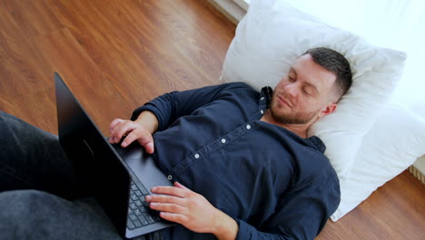 Comfortable work from home remote online. Man lies on the floor leaning on pillow and working on laptop standing on his belly. Top view.