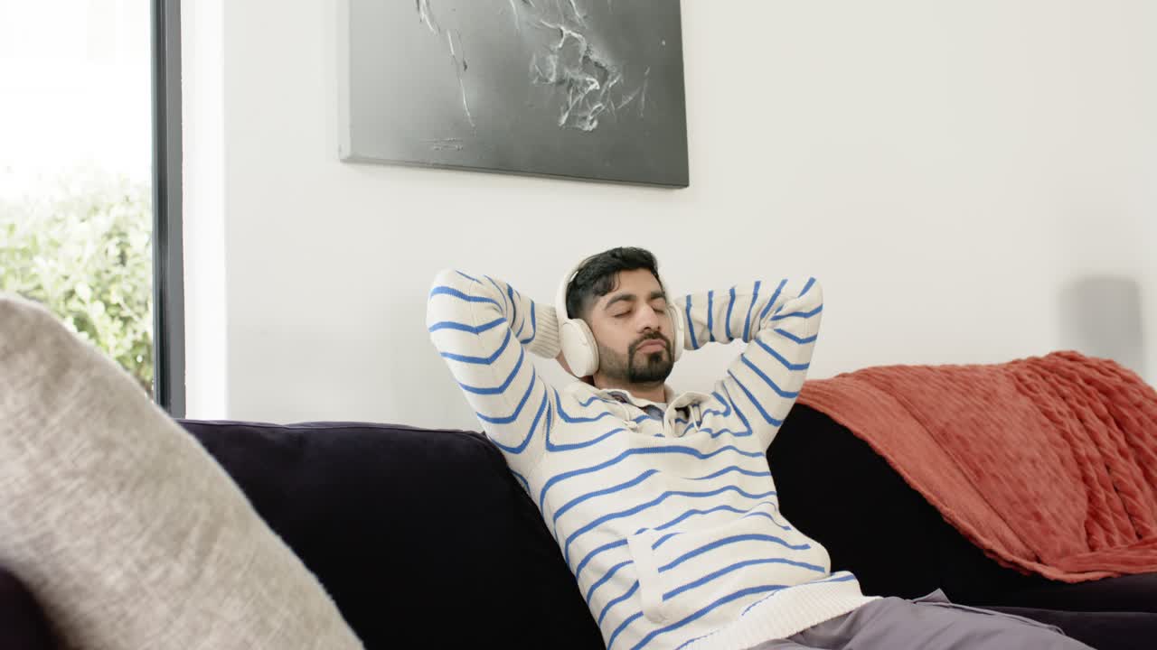 Uomo indiano con le cuffie, appoggiato allo schienale del divano di casa, che ascolta musica per rilassarsi