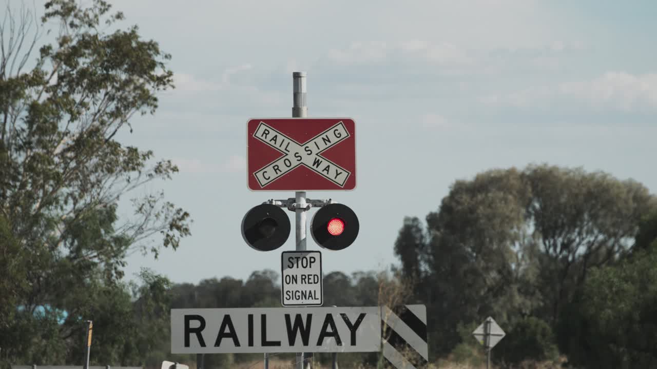 Close-up of an active railway crossing signal in rural Australia. The red light is blinking, warning of an approaching train, while signage reads “RAILWAY CROSSING” and “STOP ON RED SIGNAL.”