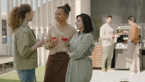 un agradable grupo multicultural de tres chicas charlando alegremente y bebiendo vino en una barbacoa en la terraza de una casa