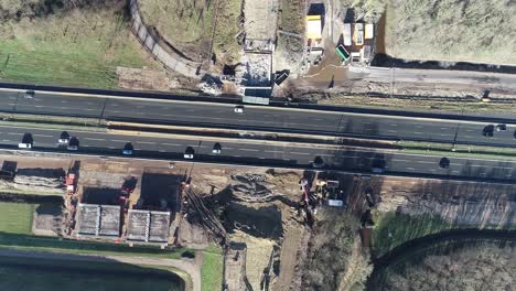 Bird's-eye-drone-shot-from-above,-you-can-see-a-highway-with-cars-and-trucks-driving,-and-a-construction-site-next-to-the-highway