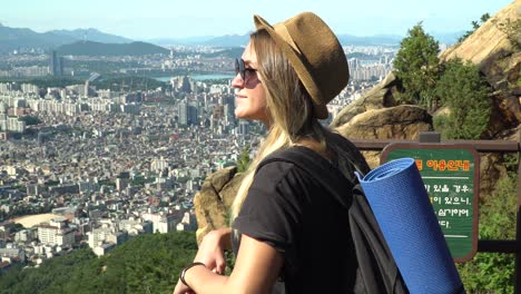 stylish blonde girl with hat and sunglasses leaning on the fence and admiring the beautiful city of seocho-gu from the gwanaksan trail on a sunny day in seoul, south korea