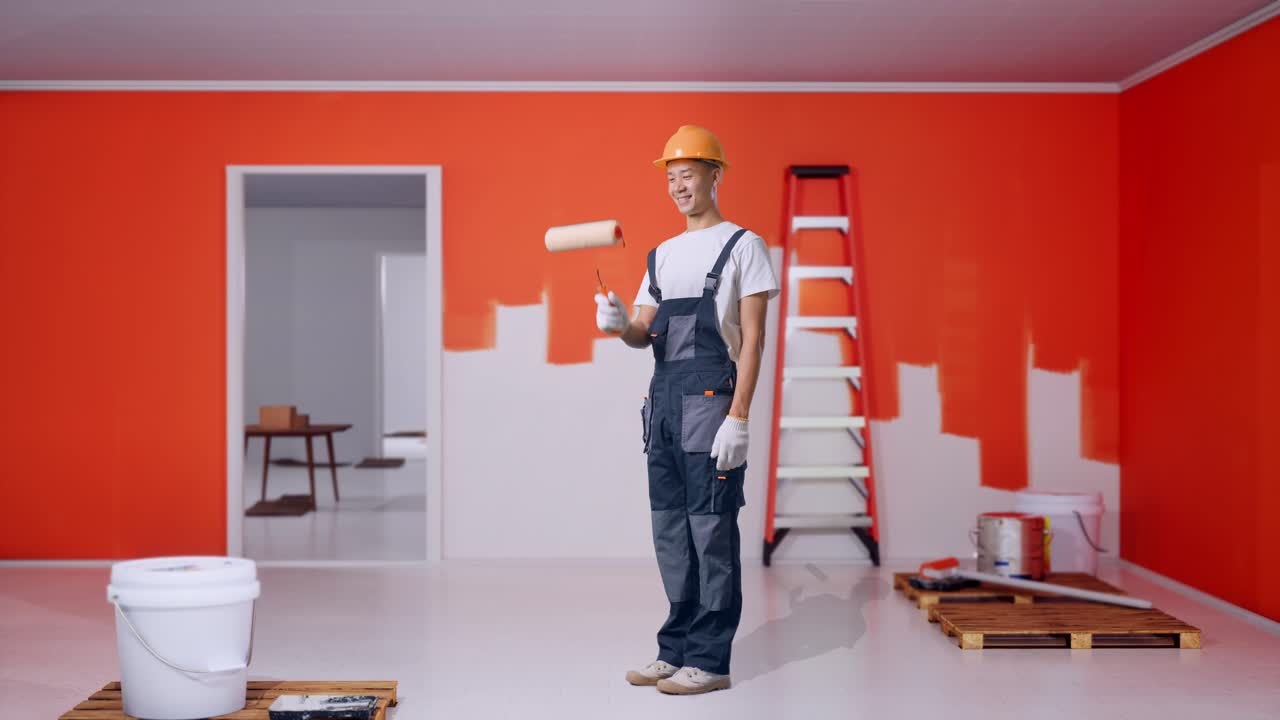 cuerpo lleno de vista lateral pintor hombre asiático con casco de seguridad pintura y sonriendo con pintura pared en la habitación