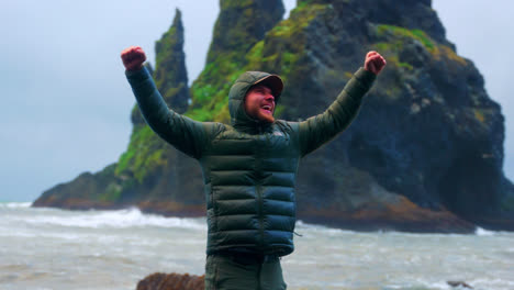 happy male tourist in winter jacket with hood raising arms standing on coastal rock at reynisfjara, southern iceland