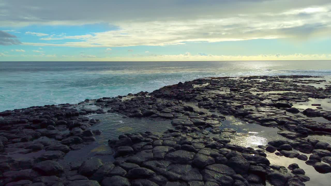 4K Hawaii Kauai boom up and pan left from behind lava rock to backlit sunlight sparkles on ocean in distance and reflects in pool on lava rocks as waves build with partly cloudy sky