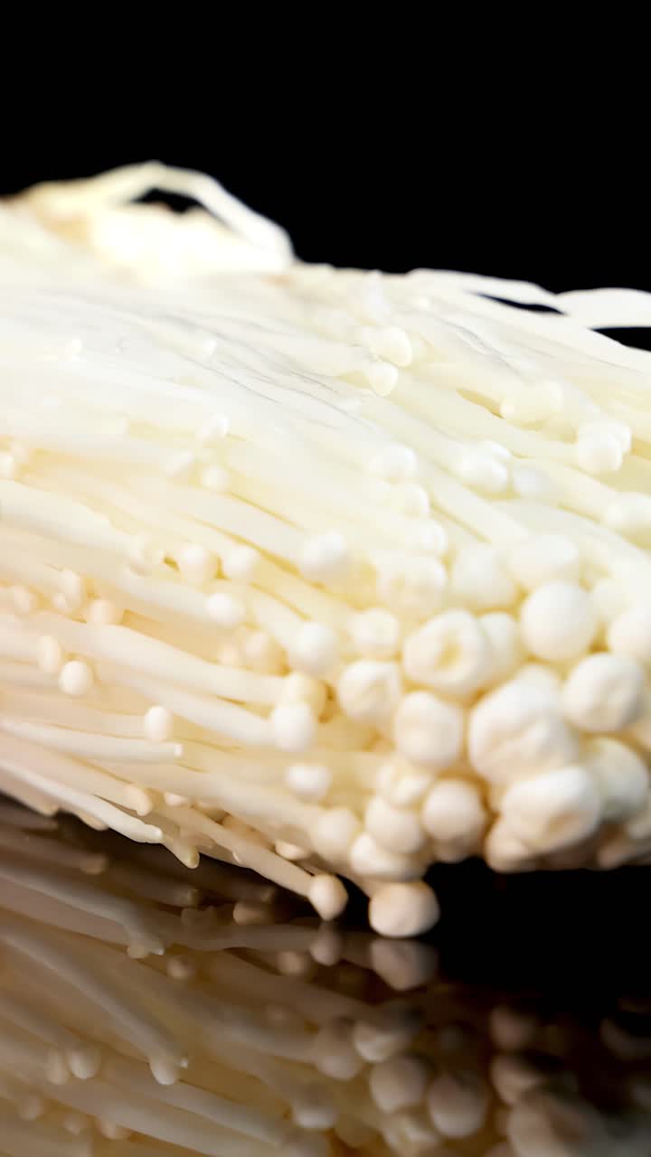 A bundle of enoki mushrooms slowly rotates on a glossy black surface under bright studio lighting, highlighting delicate textures and smooth reflections in high detail