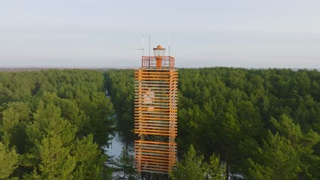 aerial view of bernati lighthouse surrounded by lush green pine tree forest with light snow, nordic woodland, baltic sea coast, sunny winter day, latvia, wide orbiting drone shot