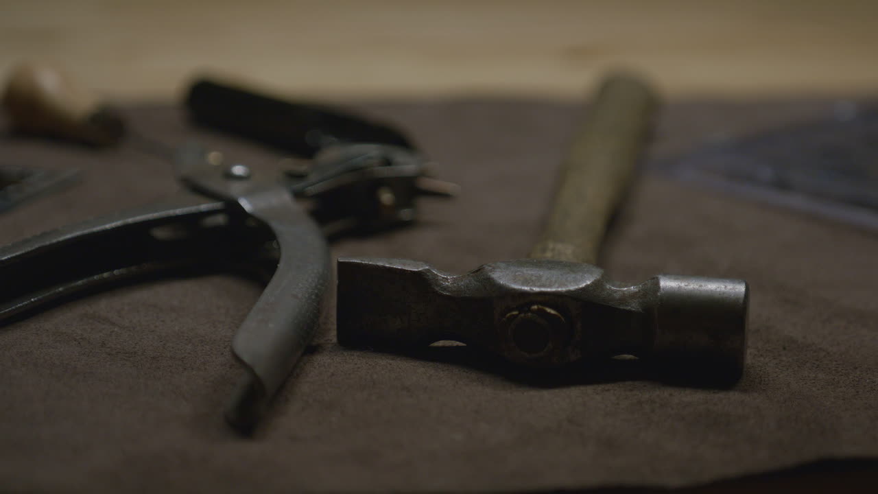 A rustic hammer with a wooden handle resting on a brown surface alongside leatherworking tools, including a hole punch and awl.