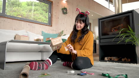 woman wrapping christmas gift at home, smiling by cozy fireplace