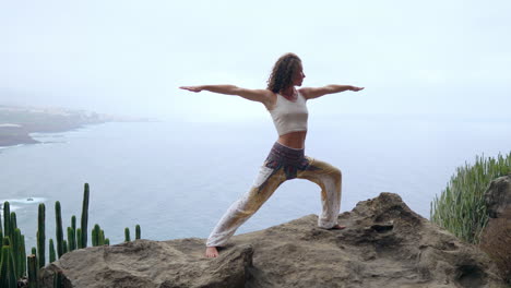 engaged in the yoga warrior pose by the ocean, beach, and rocky mountains, the woman epitomizes motivation, inspiration, and a health-conscious lifestyle