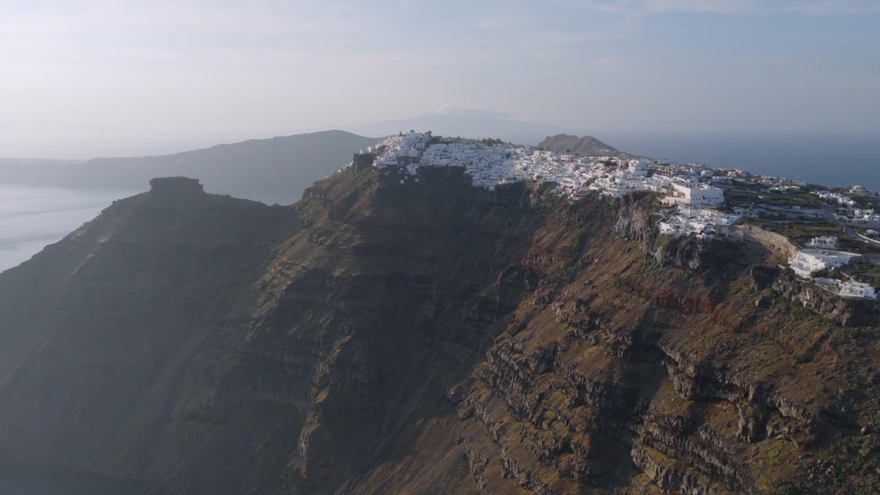 Vista aerea con drone della città di Santorini in cima alla scogliera, aggrappata al bordo della caldera, con un aspro terreno vulcanico e il mare che si estende nella morbida luce del mattino.
