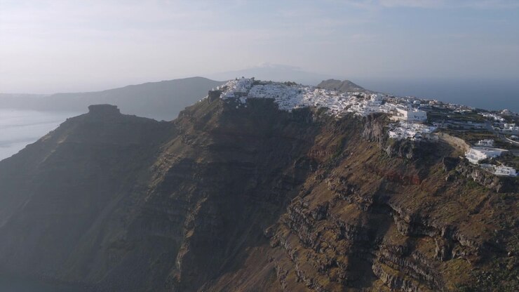 Vista aerea con drone della città di Santorini in cima alla scogliera, aggrappata al bordo della caldera, con un aspro terreno vulcanico e il mare che si estende nella morbida luce del mattino.