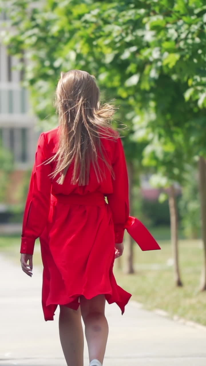 Back view of a woman in summer. Young female in red dress with long hair is walking on the street to the building in the city.Slow motion. Vertical video
