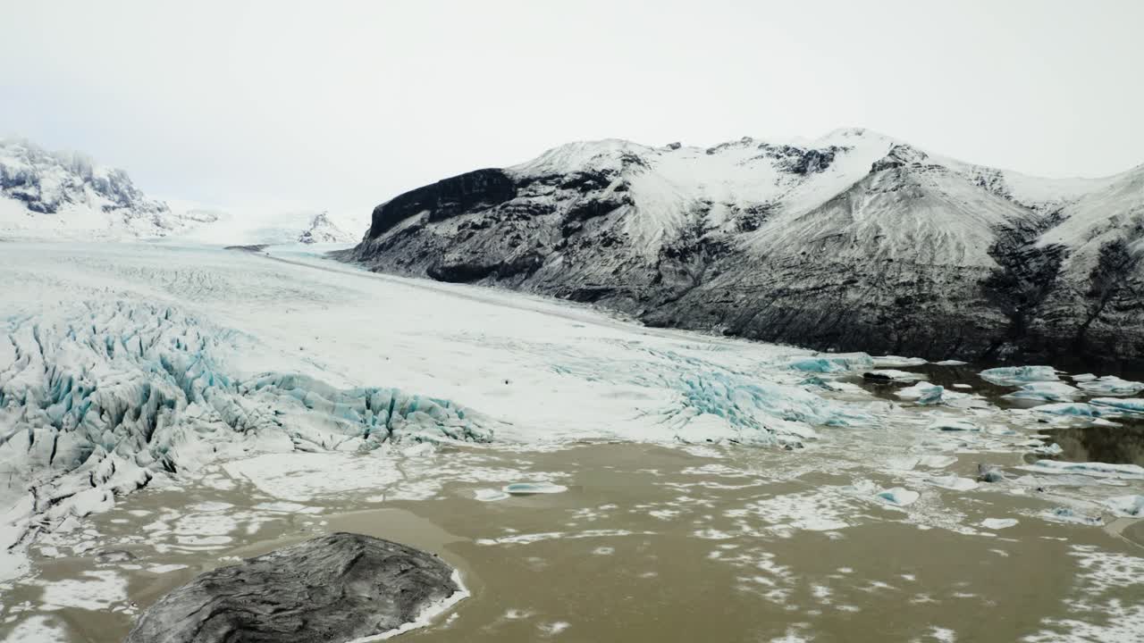 Drone footage capturing the intricate ice formations of Svinafellsjokull glacier blending seamlessly with the surrounding muddy lagoon and rocky terrain.