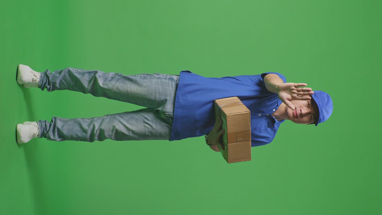 cuerpo lleno de mensajero masculino asiático en uniforme azul haciendo un gesto de parada y sacudiendo la cabeza mientras entrega una caja de cartón en el fondo de la pantalla verde en el estudio
