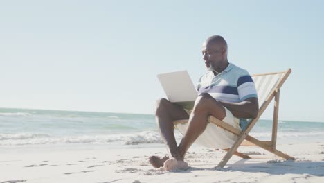 Senior-african-american-man-on-deck-chair,-using-laptop-at-beach,-copy-space,-in-slow-motion
