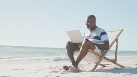 Hombre-Afroamericano-De-Alto-Rango-En-Una-Tumbona,-Usando-Una-Computadora-Portátil-En-La-Playa,-Copiando-Espacio,-En-Cámara-Lenta