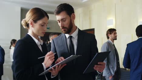 Caucasian-businesswoman-reading-documents-and-charts-and-her-male-colleague-holding-a-tablet-while-they-talking-and-discussing-something-in-a-conference-hall