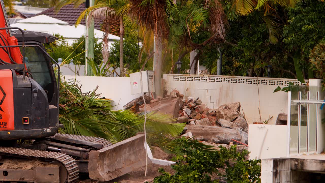 An orange excavator removes a large palm tree from a residential garden in daylight, with lush greenery and a white fence in the background. Camera remains steady