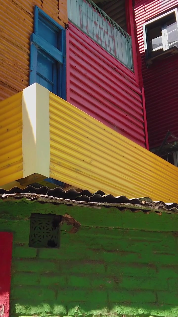 POV caminhando por fachadas coloridas de metal corrugado em amarelo, vermelho, verde e azul com detalhes de telhado de telha em Caminito, La Boca, Buenos Aires, Argentina.