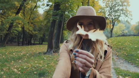 rear view of caucasian young blonde woman in a hat and coat walking in the park and leading the camera with her, then she turns and smiles while holding a yellow leaf