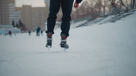 rear view skater wearing black skates skating on ice surface with visible blade marks and snow blurred trees and other skaters in background outdoor winter rink under soft evening light