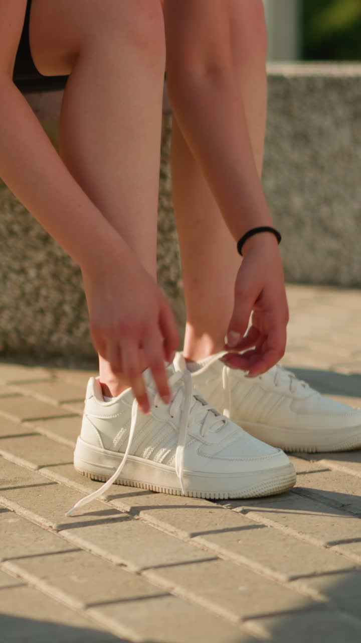 Partial view of lady seated on bench outdoors removing left sneaker with left hands,while her right hand is on the leg sunlight casting shadows on paved ground, greenery in background