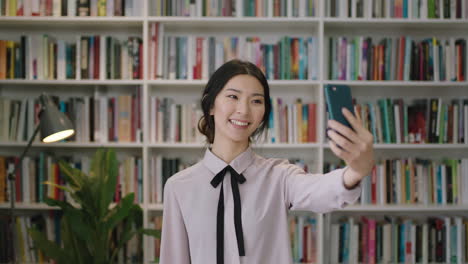 portrait beautiful asian woman standing in library using smartphone video chatting selfie bookcase in background smiling waving