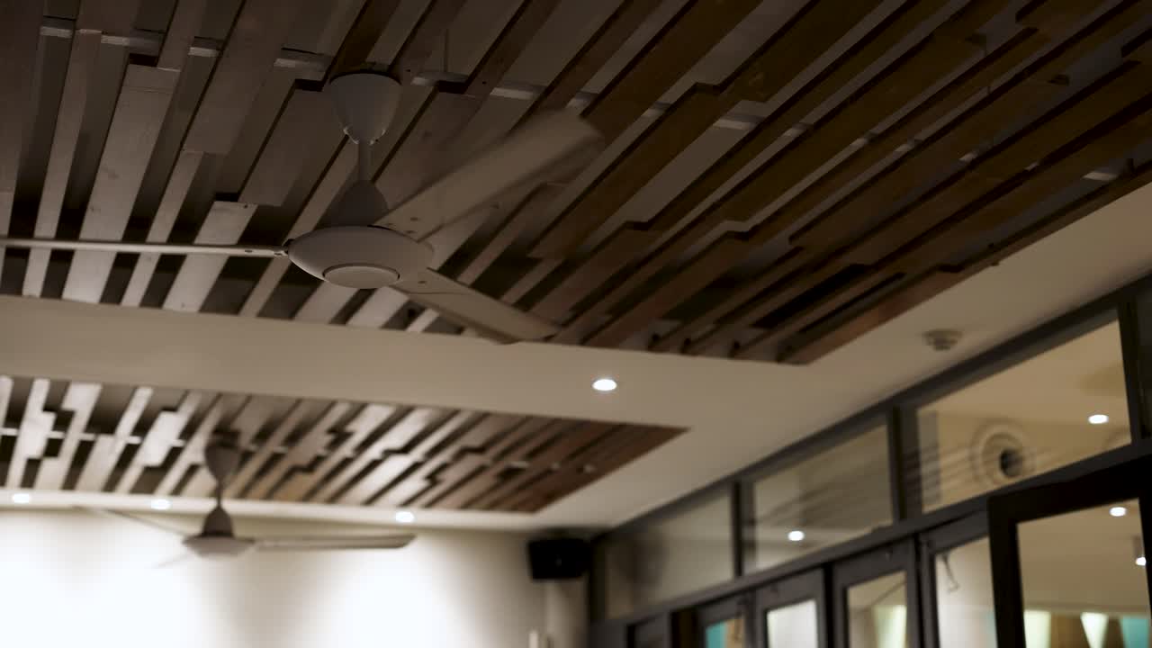 Un ventilatore a soffitto ruota in una stanza contemporanea con doghe in legno e finestre in vetro, creando un'atmosfera calma e ariosa.