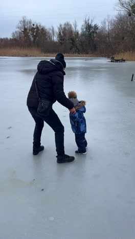 dad and son are on the winter walk on the frozen river. father supports the baby sliding by the ice. vertical video.